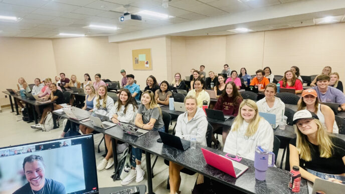 Classroom full of students smiling with J.D. Tuminski on a computer monitor in the foreground.