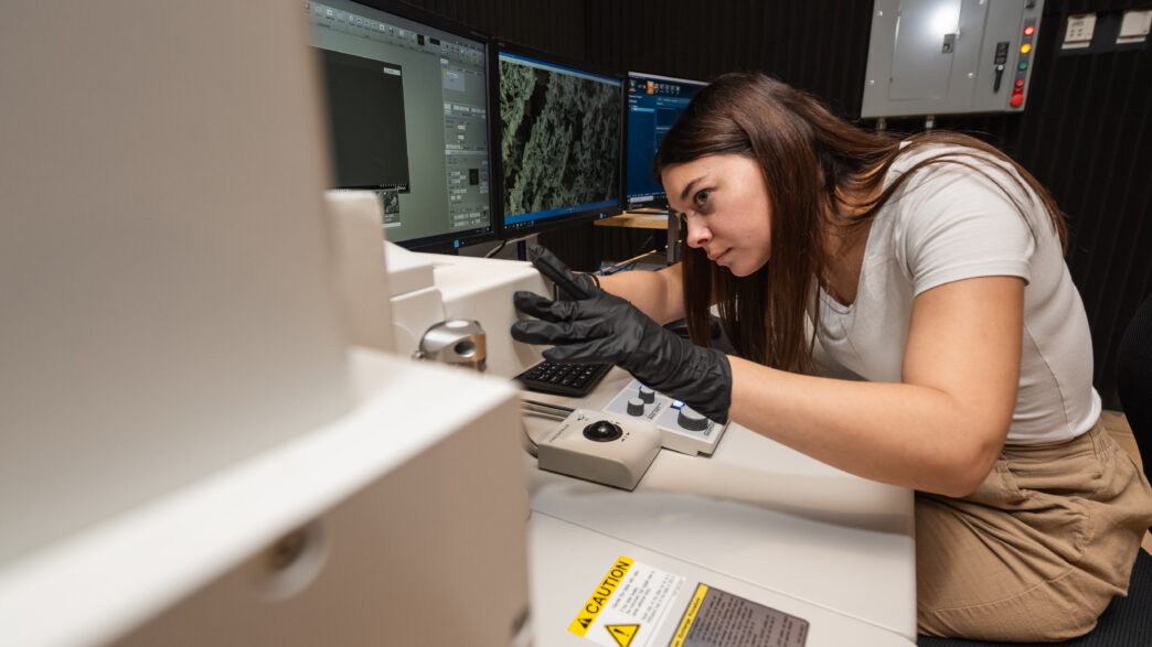 A young woman is at work in a lab with computers and wears black, latex gloves.