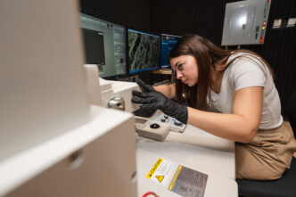 A young woman is at work in a lab with computers and wears black, latex gloves.