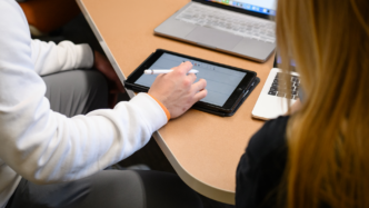 Two people sit at a table during an interview, with one person taking notes on a tablet beside open laptops.