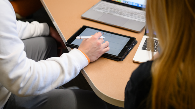 Two people sit at a table during an interview, with one person taking notes on a tablet beside open laptops.