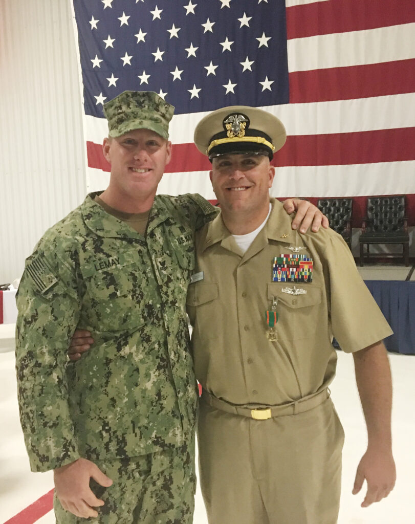 A Naval officer and a sailor pose together with an American flag behind them