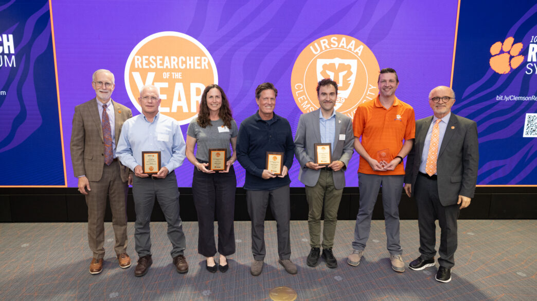 A group of seven people stand side by side in front of a large screen with the Researcher of the Year logo on it.