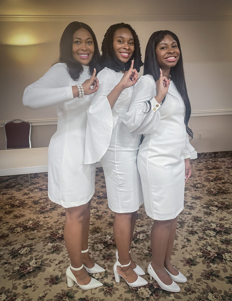 Three African American women stand close together with their shoulders to the camera and display hand signals