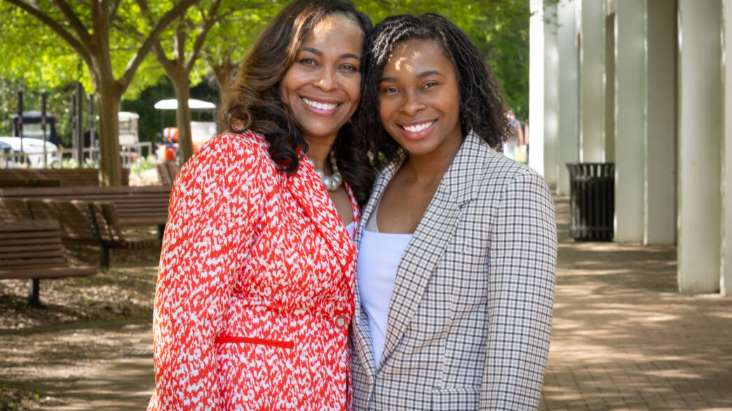 Two African American women in business suits stand in the shade of some trees next to Cooper Library and smile at the camera with their arms around each other