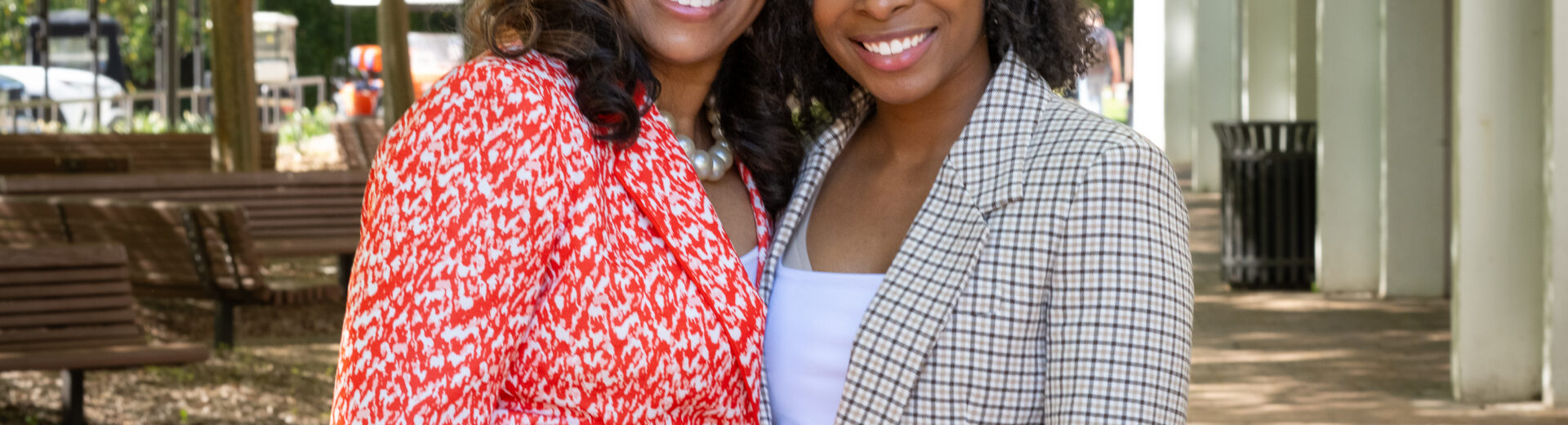 Two African American women in business suits stand in the shade of some trees next to Cooper Library and smile at the camera with their arms around each other