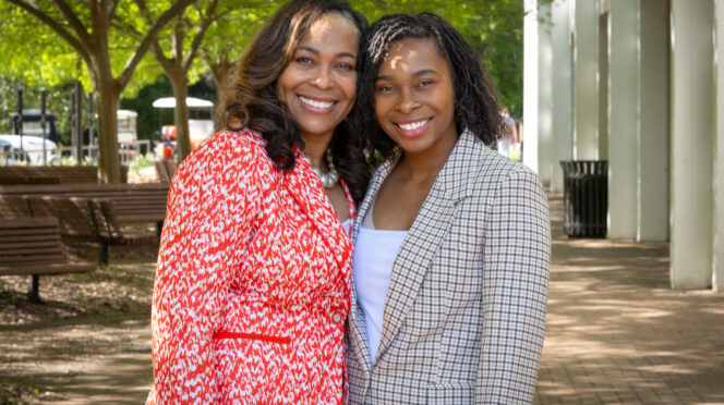 Two African American women in business suits stand in the shade of some trees next to Cooper Library and smile at the camera with their arms around each other