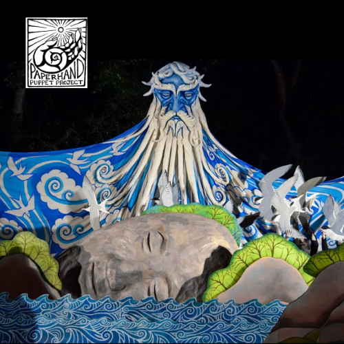 A large puppet resembling an ocean deity is surrounded by white bird puppets, alongside a large gray rock puppet with a carved face.