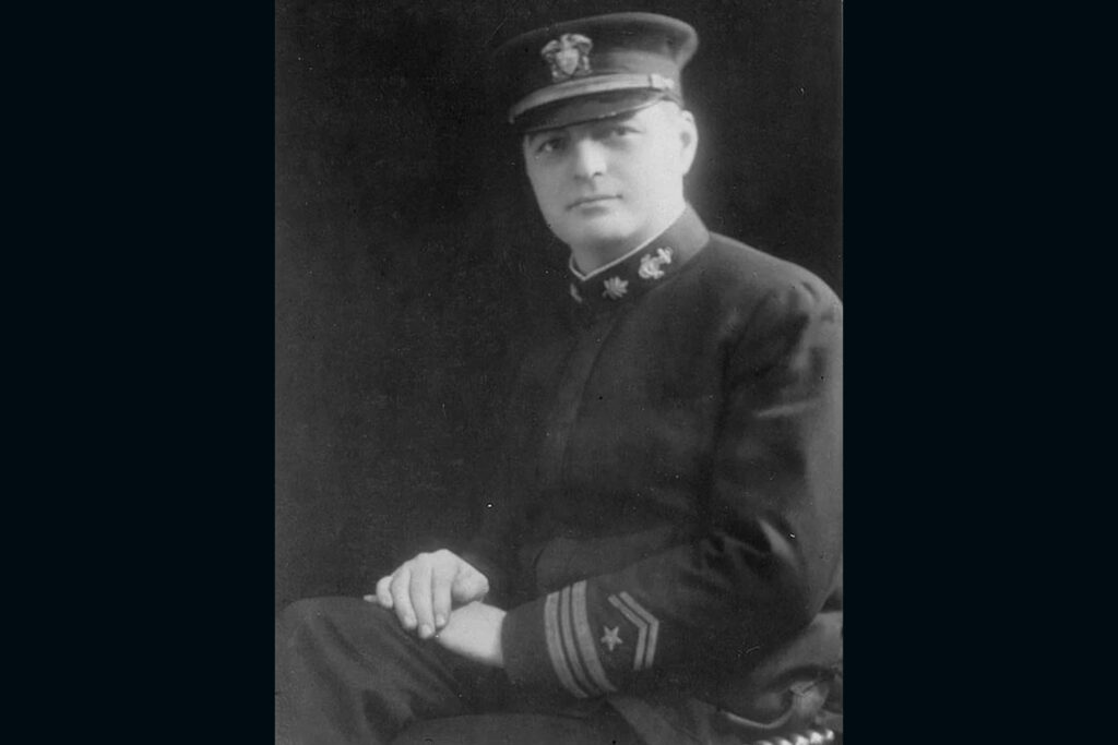Archival black and white formal photograph of a young Navy officer in formal uniform, sitting with his hands on one leg and looking over his left shoulder at the camera