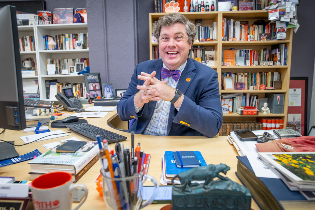 A man sits at a cluttered desk smiling with his elbows on the desk and his hands clasped