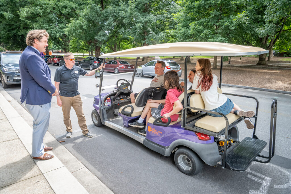 A man stands next to a golf cart chatting with the four people in it