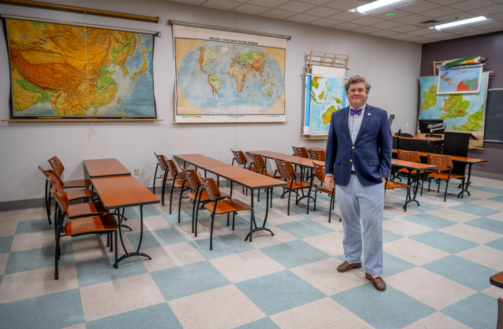 A man stands on the tiled floor of a classroom in front of a row of wood desks and a wall covered in large maps.