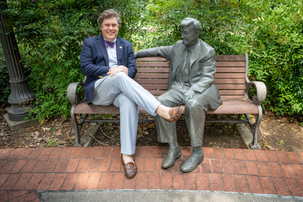 A man sits on a bench next to a bronze statue of another man sitting on the same bench