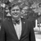 Black and white photograph of a man wearing a suit jacket and bowtie standing in a park behind Tillman Hall