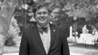 Black and white photograph of a man wearing a suit jacket and bowtie standing in a park behind Tillman Hall