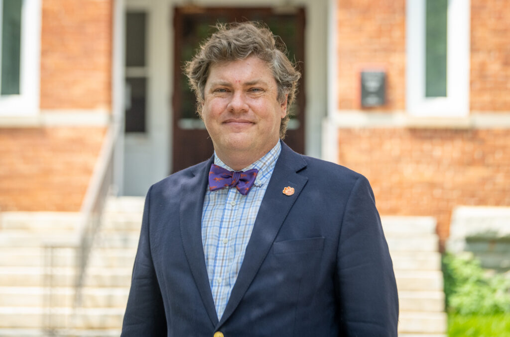 A man in a jacket and bowtie stands in front of a brick building