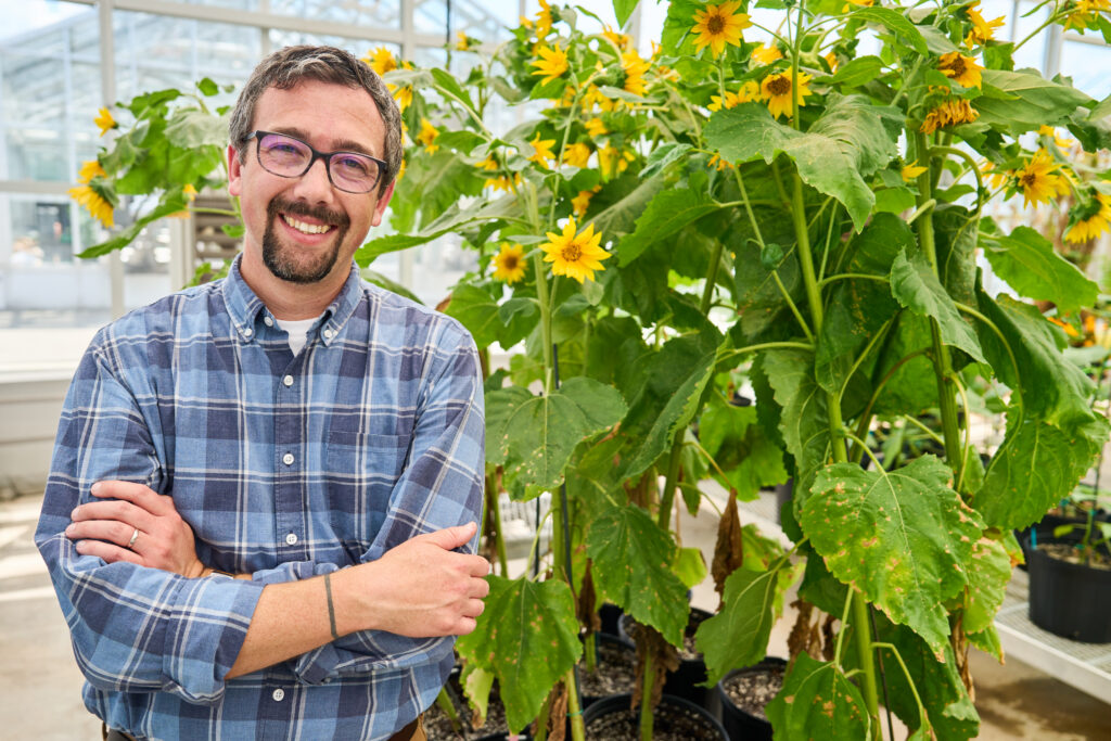A man wearing a blue plaid shirt with dark eyeglasses with a mustache and goatee stands in front of a sunflower plant.