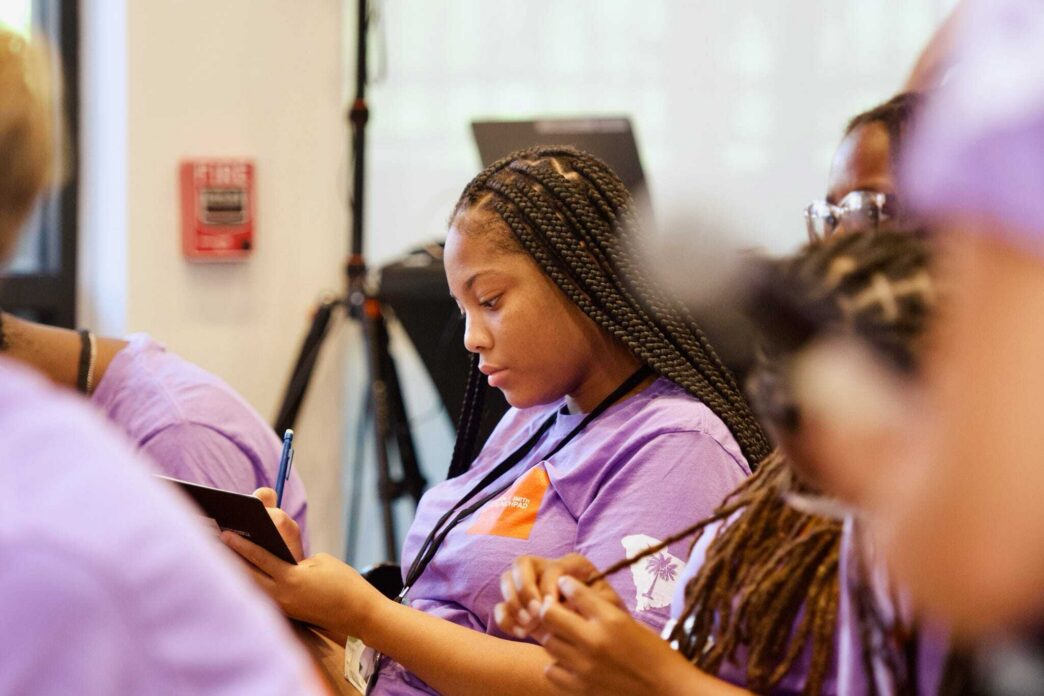A young Black woman wearing a purple shirt and blue lanyard takes notes in a classroom environment.