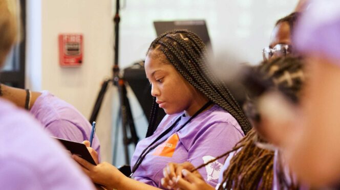 A young Black woman wearing a purple shirt and blue lanyard takes notes in a classroom environment.