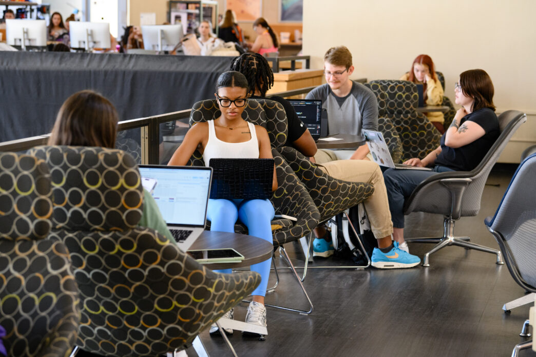 A group of students sit in chairs while using laptops in Cooper Library, with Adobe Studio in the background.