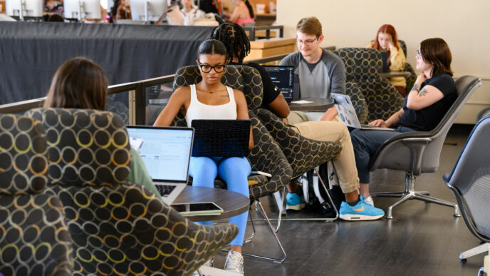 A group of students sit in chairs while using laptops in Cooper Library, with Adobe Studio in the background.