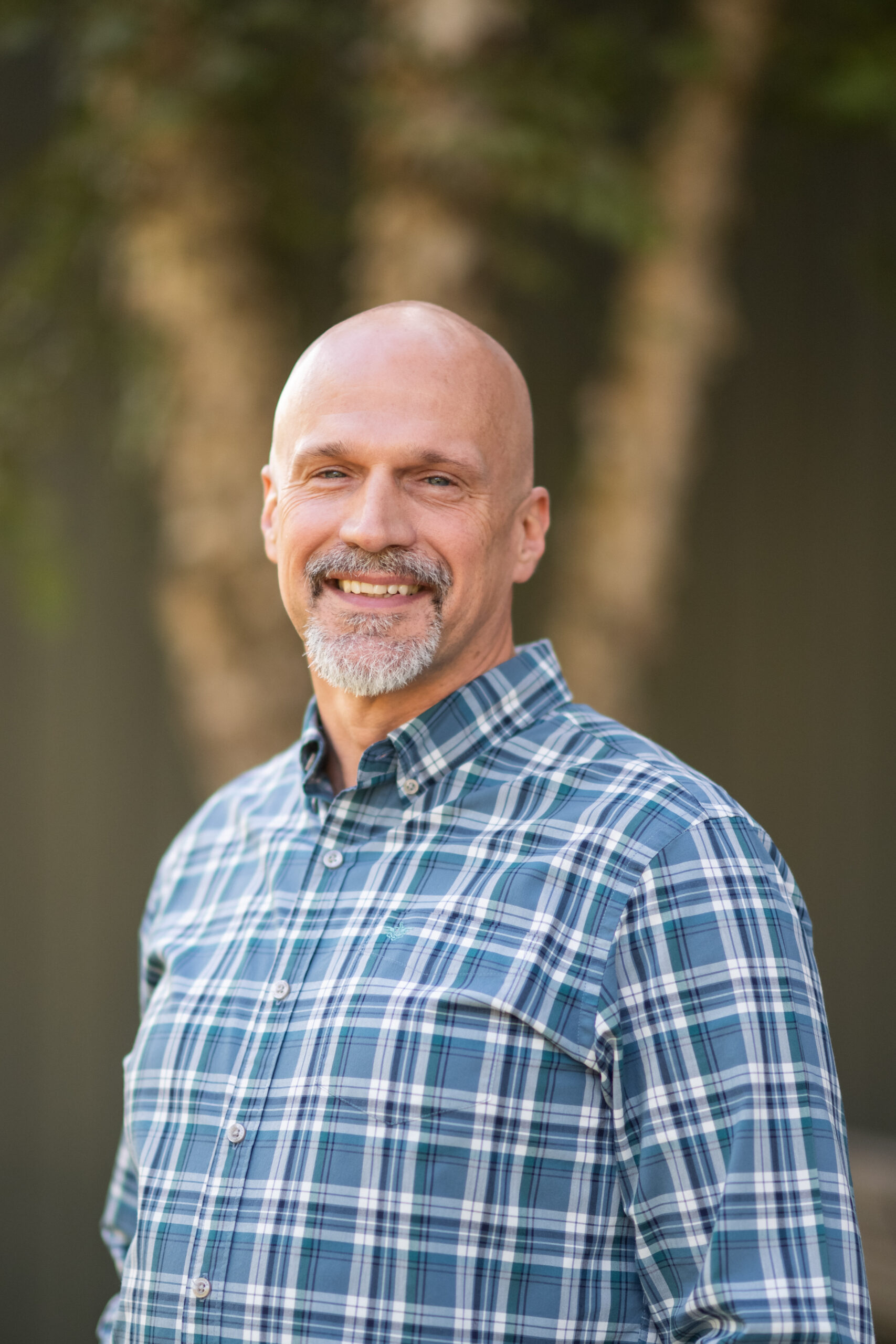 smiling bald and bearded man, standing in front of a tree