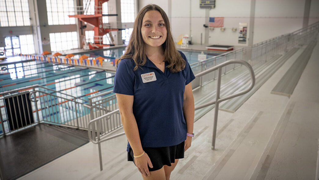 A woman stands in the bleachers above a swimming pool with the diving platform over her right shoulder and an American flag over her left.