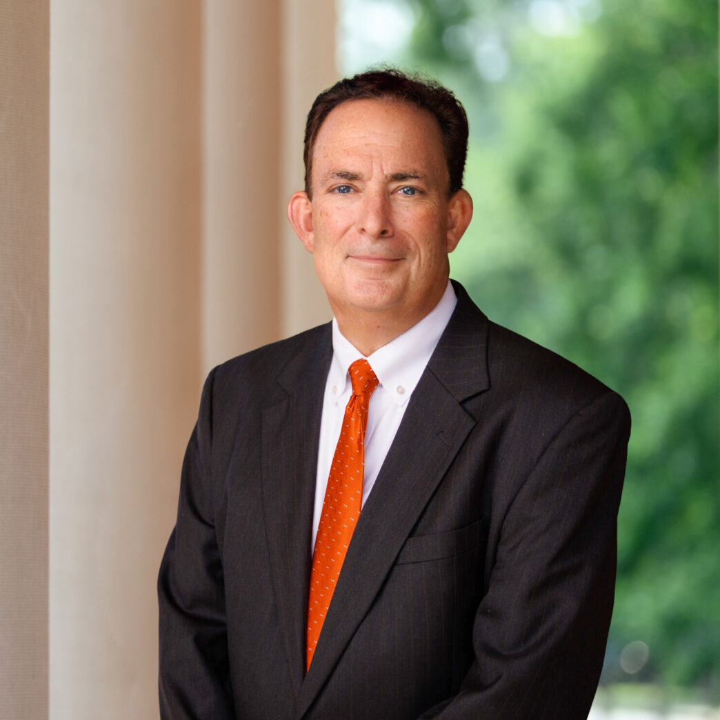 A man wearing an orange tie and grey suit stands in front of white columns outdoors.