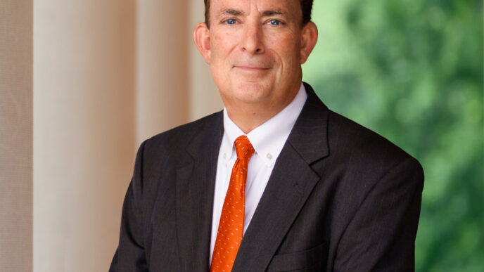 A man wearing an orange tie and grey suit stands in front of white columns outdoors.