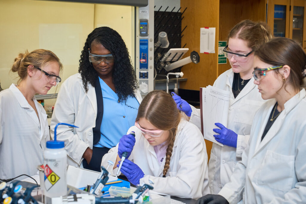 A Black woman with a blue shirt and wearing a lab coat watches a student pipette while three other students in white lab coats watch.