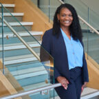 A Black woman with long black hair wearing a black suit and blue shirt stands on a stairway with her hand on the rail looking at the camera.