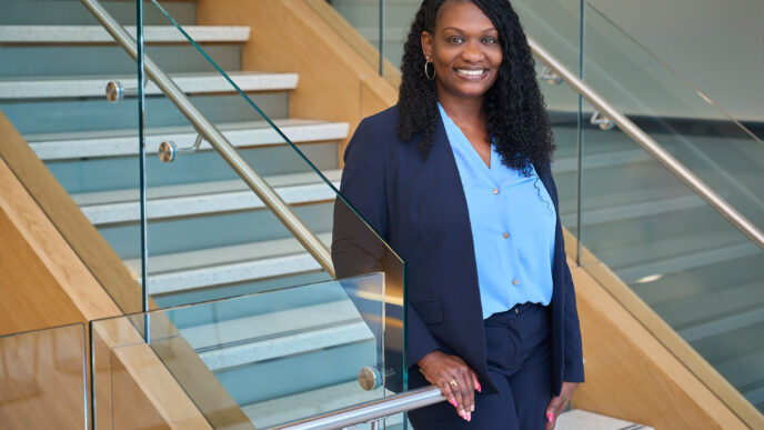 A Black woman with long black hair wearing a black suit and blue shirt stands on a stairway with her hand on the rail looking at the camera.