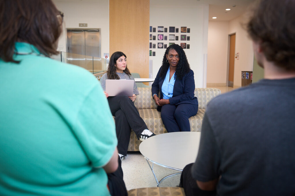 A woman wearing a blue shirt and black suit talks to students sitting on a couch.