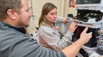 A man with a beard and goatee wearing a gray sweatshirt points at a machine in a chemistry lab while a woman watches.