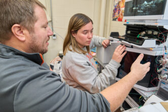 A man with a beard and goatee wearing a gray sweatshirt points at a machine in a chemistry lab while a woman watches.