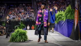 A smiling man wearing a graduation cap and gown with multiple sashes over his shoulder walks across a stage toward the camera