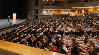 A large audience in an auditorium looks on at a speaker on a stage