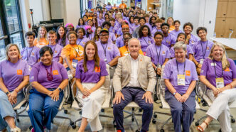 A man in a suit coat sits in the front row of a room full of people in purple T-shirts sitting in rows