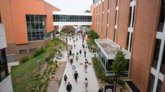 A concrete campus walkway between brick academic buildings, with dozens of students walking in both directions, landscaped gardens and bicycle racks along the path, and an enclosed pedestrian bridge connecting the buildings overhead.