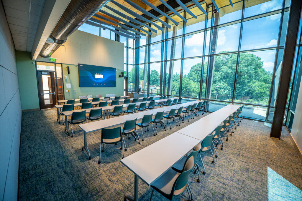 A large classroom with three long rows of tables, a screen at one end, and floor to ceiling windows looking out onto trees and blue sky making up an entire wall