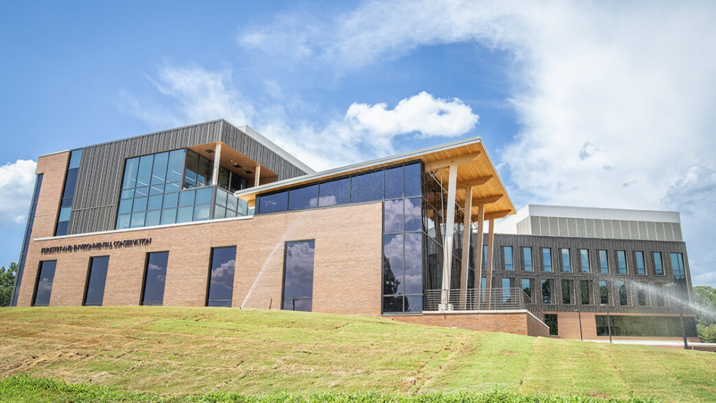 Exterior shot of a modern building with large windows and exposed wood beams under a blue sky with clouds swirling in it.