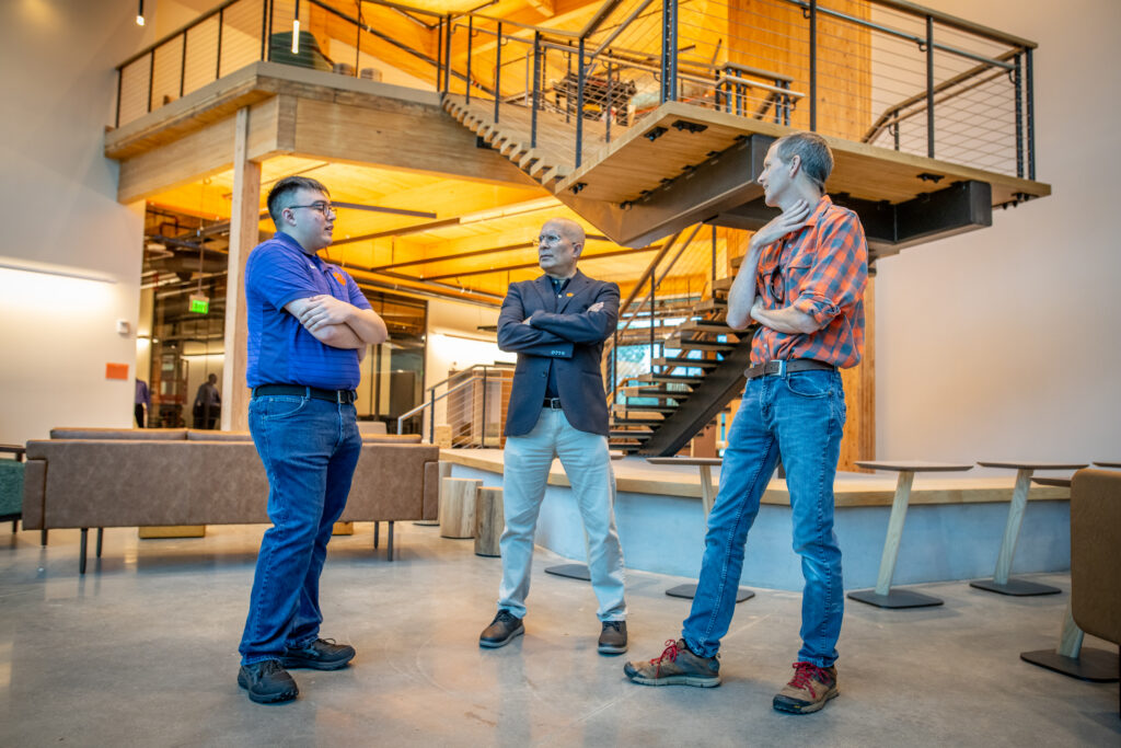 Three men stand in a large room beneath a grand staircase that switchbacks up to a second floor behind them