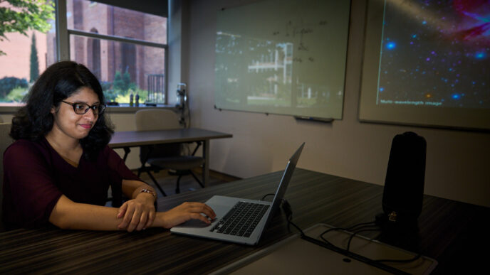 A woman with dark hair and glasses sits at a table looking at a computer with a monitor on the wall displaying a supermassive black hole.,