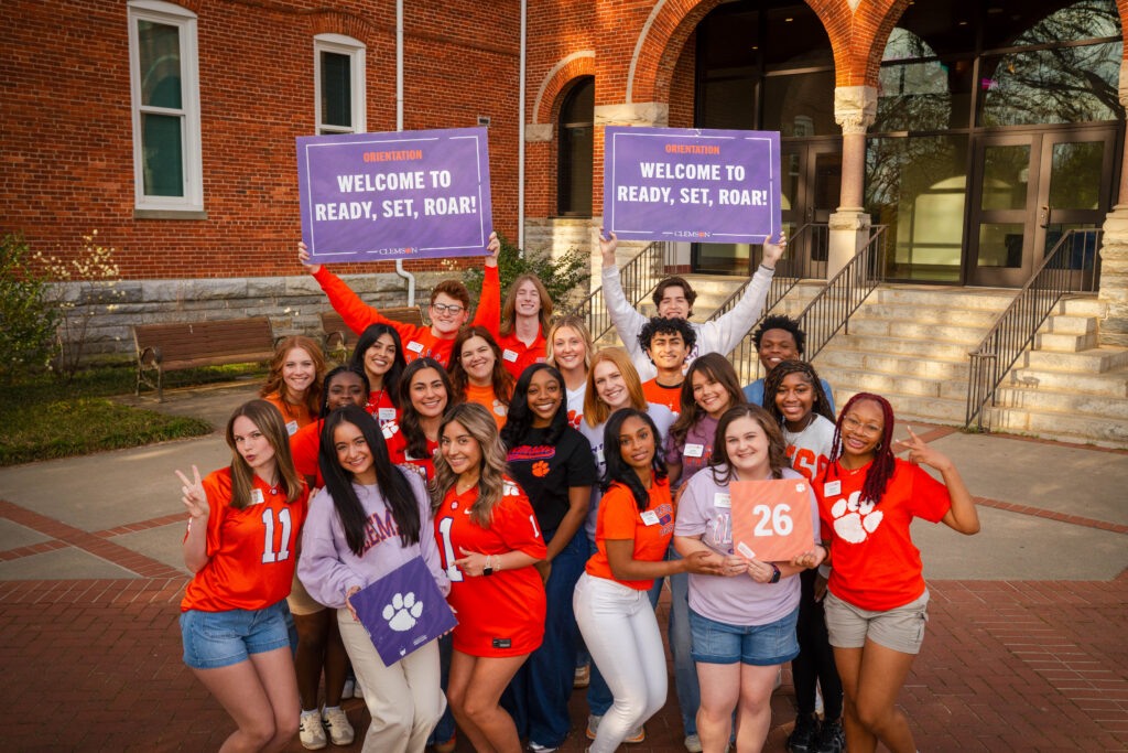 The 2026 summer orientation team stands in front of Tillman Hall posing together with "Welcome to Ready, Set, Roar!" signs.