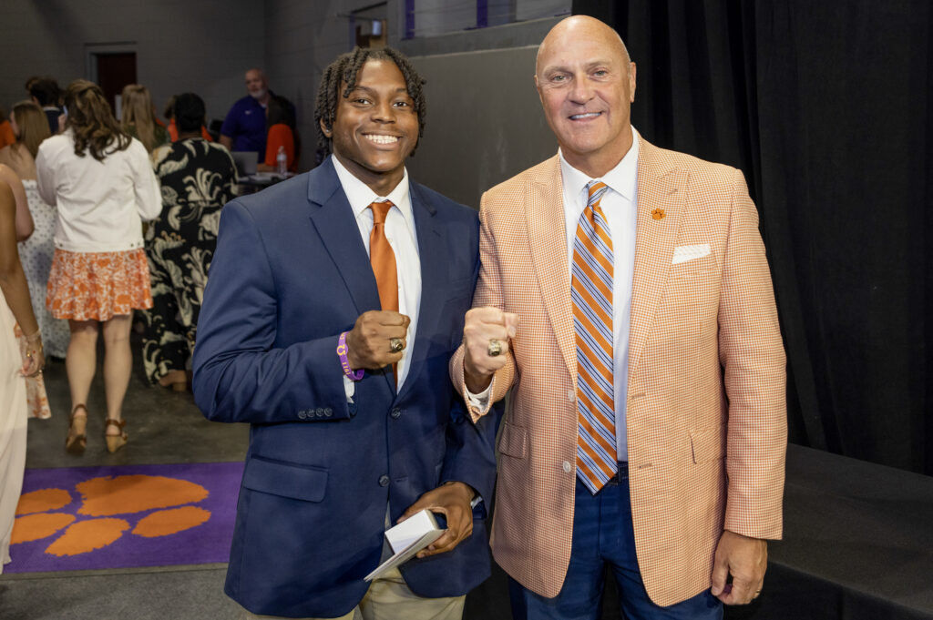 Two men in suits stand next to each other making fists and smiling at the camera