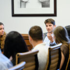 A man in a suit speaks while sitting at a table, surrounded by others