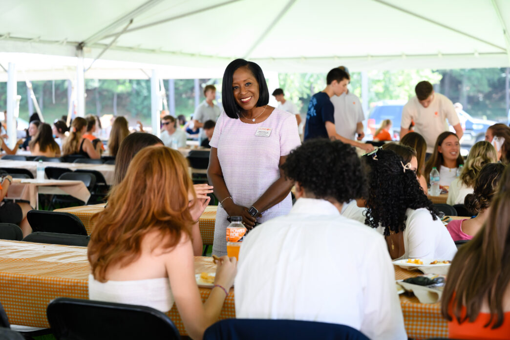 A female in a dress speaks to individuals seated at a table