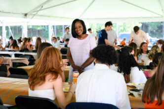 A female in a dress speaks to individuals seated at a table