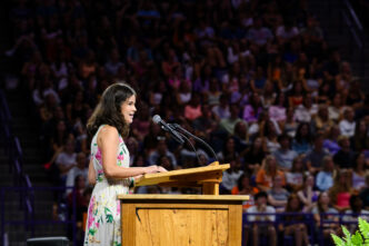 A female in a dress speaks from behind a podium to a large crowd
