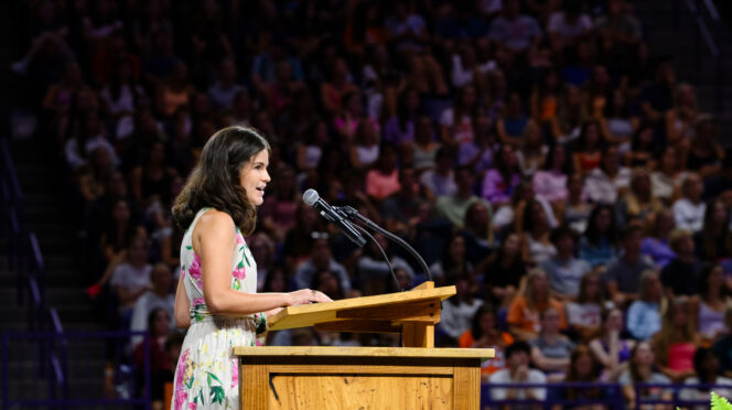 A female in a dress speaks from behind a podium to a large crowd